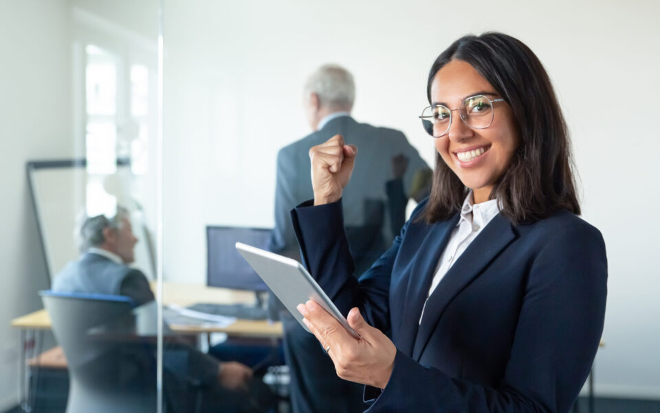 Happy female professional in glasses and suit holding tablet and making winner gesture while two businessmen working behind glass wall. Copy space. Communication concept